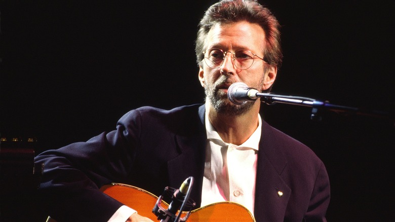 Eric Clapton sitting down on stage, playing an acoustic guitar and singing into a microphone