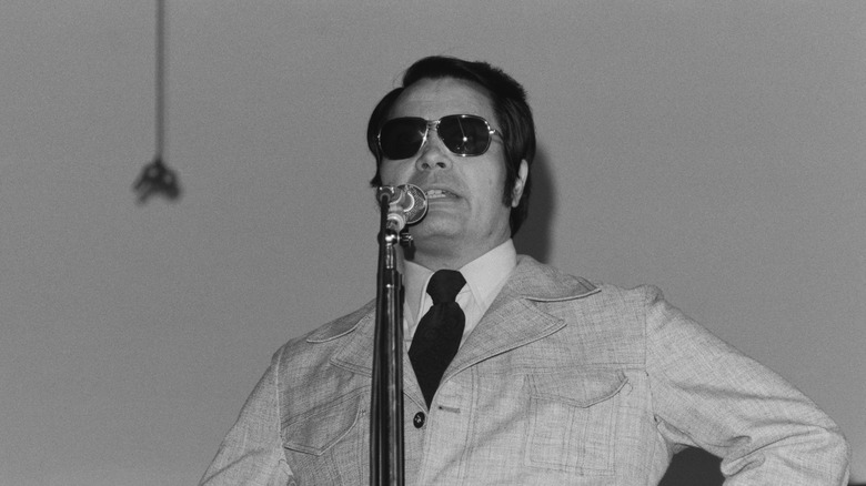 Jim Jones wearing aviators speaking at a microphone