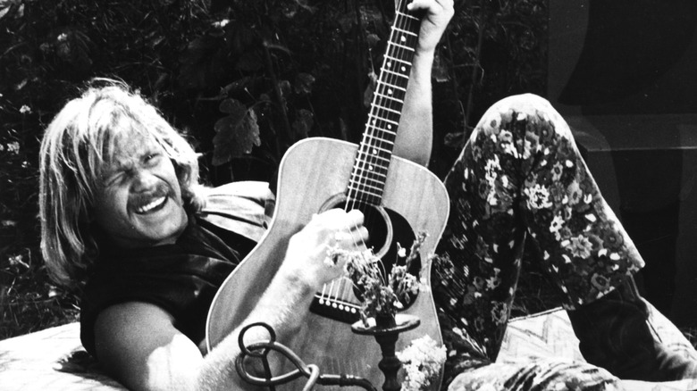 Barry McGuire smiling while playing guitar laying down outside