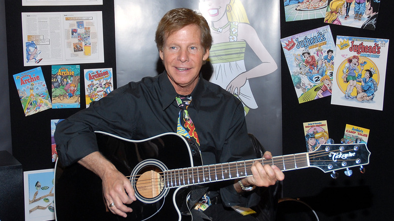 Ron Dante of the Archies playing a guitar in front of a poster of several covers of Archie comics