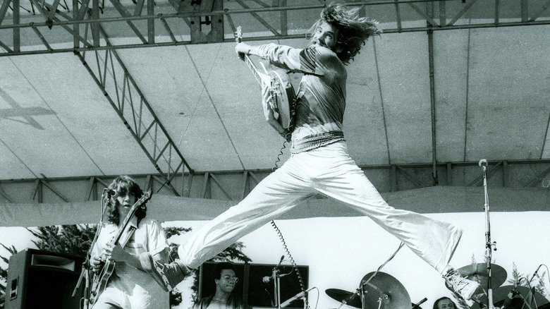 Kenny Loggins on stage, jumping in the air while playing guitar.
