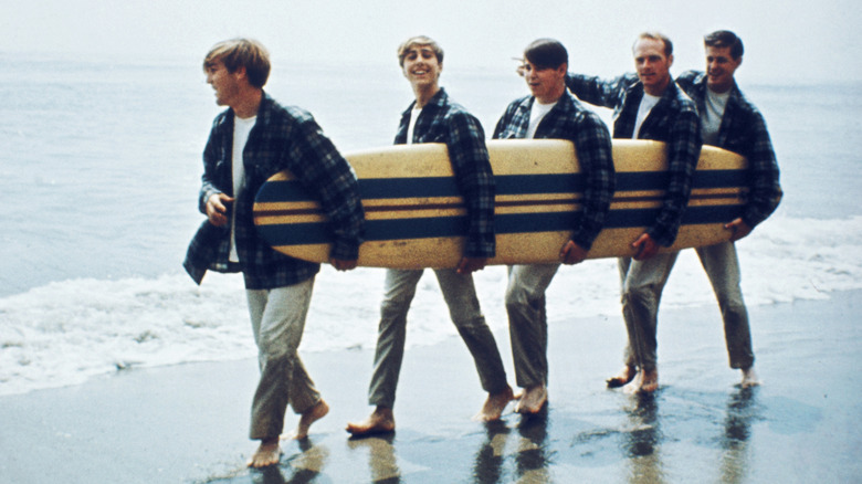 The Beach Boys on a beach, carrying a long surfboard