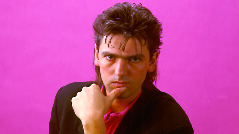 Robyn Hitchcock in dark suit with thumb on chin with pink background