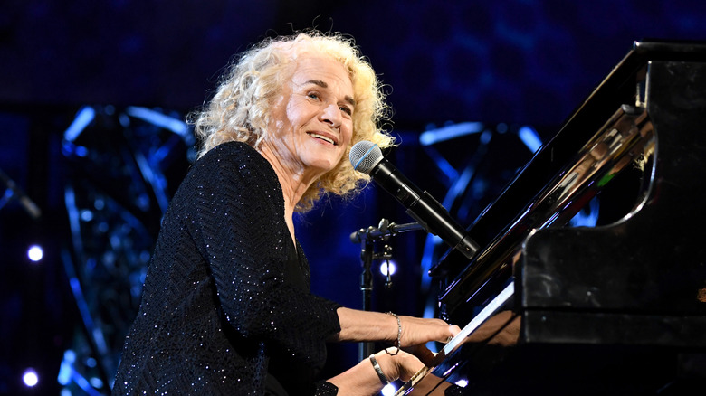 Carole King smiling and playing piano onstage