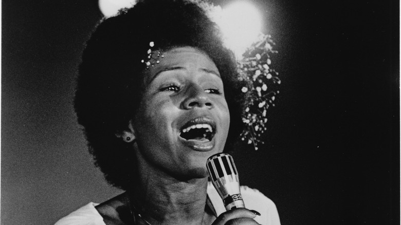 Black and white photo of Minnie Riperton holding a microphone with flowers in her hair performing on television in 1975