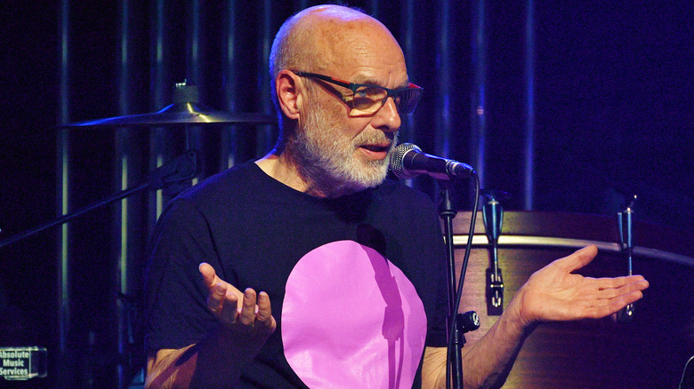 Brian Eno gesturing with both hands speaking into a microphone onstage