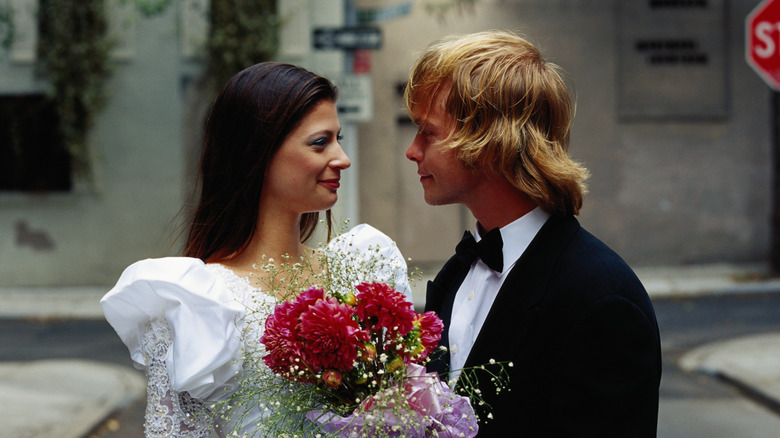 Couple in 1980s-styled wedding dress and tuxedo look at one another.