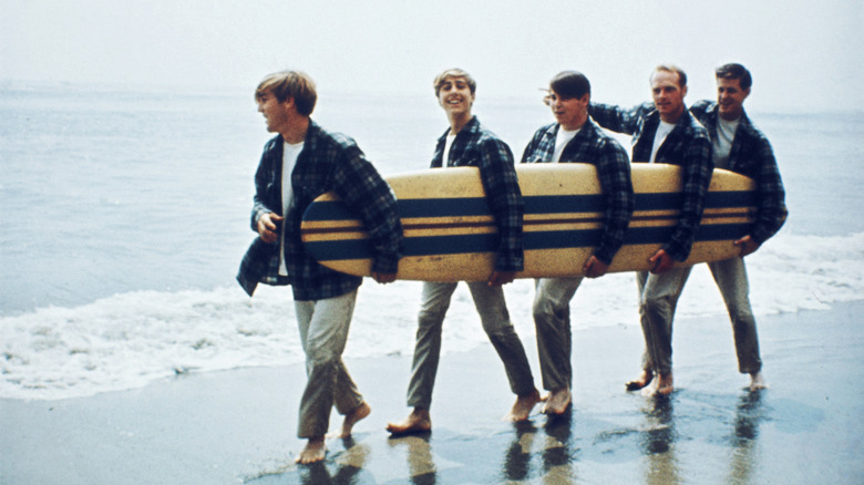 The Beach Boys carrying a surfboard at the edge of the ocean