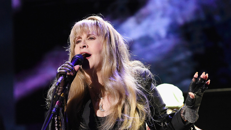 Stevie Nicks singing into a microphone on stage and gesturing.