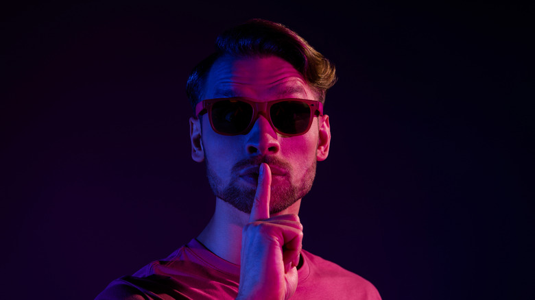 Man in 1980s sunglasses with making silence gesture with finger to mouth