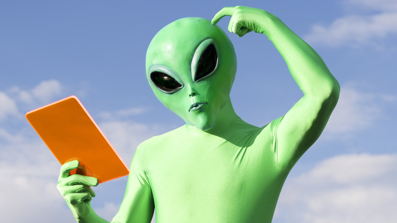 A green alien looking confused at an orange tablet