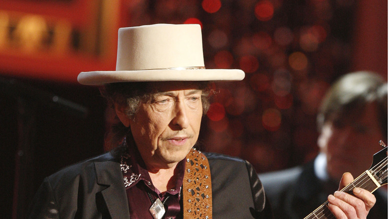Bob Dylan holding a guitar on stage while wearing a hat