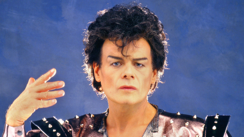 Garry Glitter in '70s glam dress holding up hand with blue background