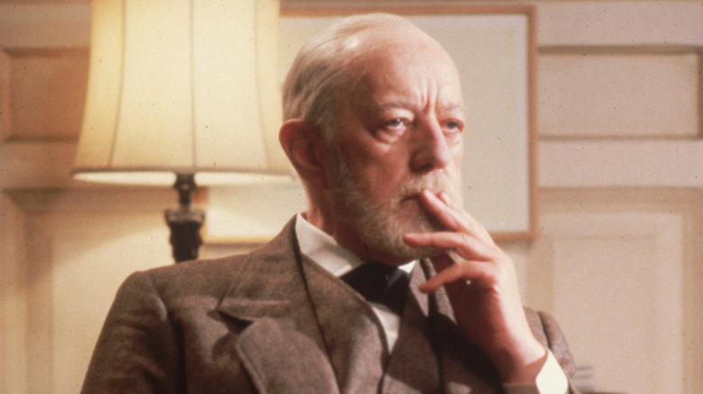 Alec Guinness sat in grey suit with white beard looking thoughtful