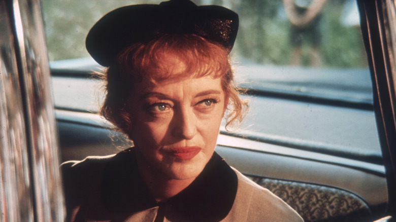 Bette Davis sat in car wearing light jacket and a hat