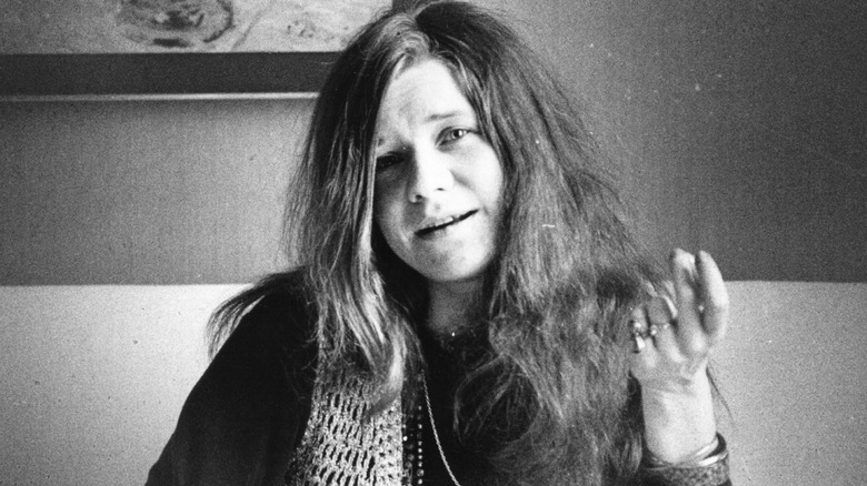 Janis Joplin sat smiling on sofa with hand raised