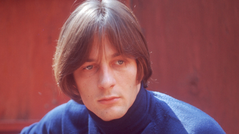 Photo of Gene Clark from around 1970 wearing a blue sweater against a reddish wooden background, playing an unpictured guitar