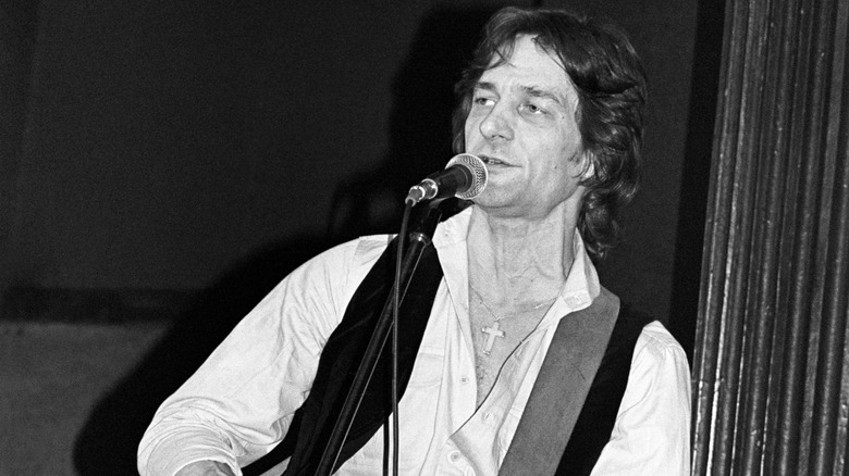 Black and white photo of Gene Clark performing on stage in 1979 wearing a cross necklace, white button up, vest, and standing in front of a microphone stand