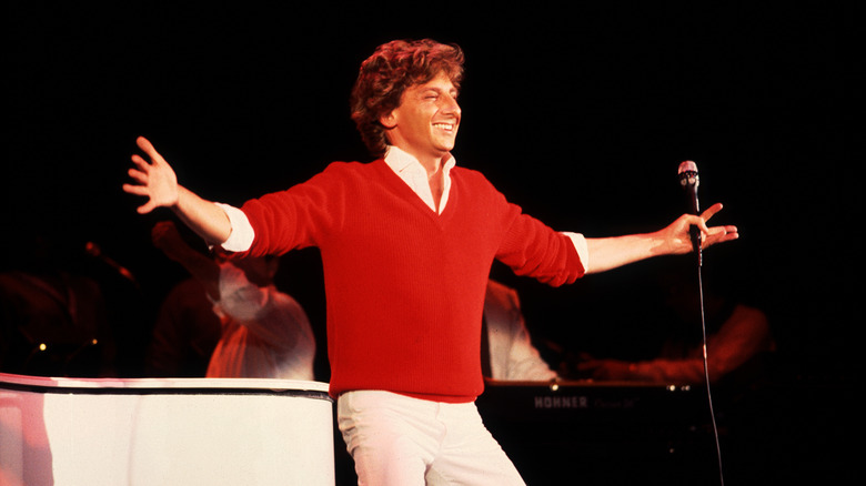 Barry Manilow smiling with arms out wearing red cardigan on stage in 1982
