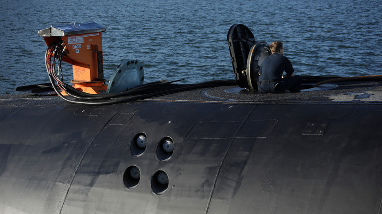 A member of a submarine crew sitting on near an open hatch on top of the sub looking out at the water