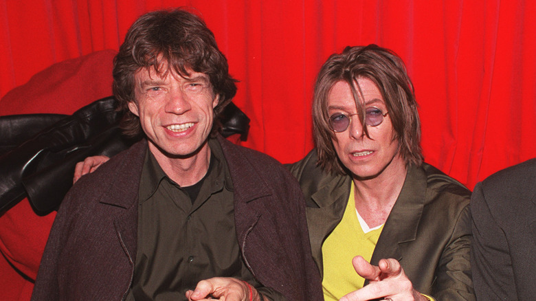 Mick Jagger seated next to David Bowie, in front of red curtains