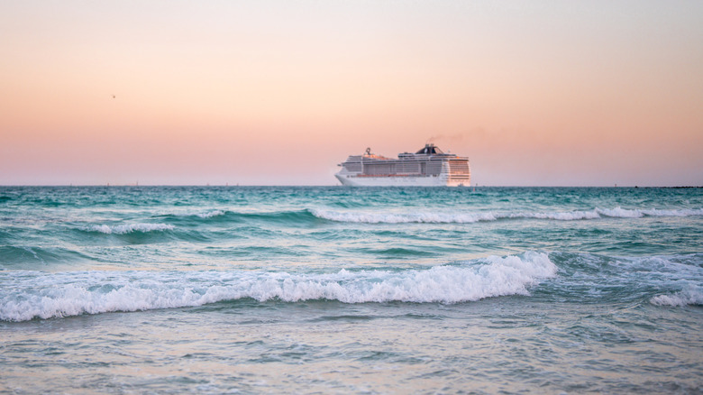 A white cruise ship sails on the ocean at sunset while waves hit the sandy shore.