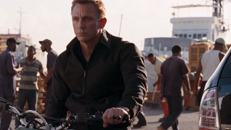 daniel craig on motorcycle in foreground as extra sweeps air in background
