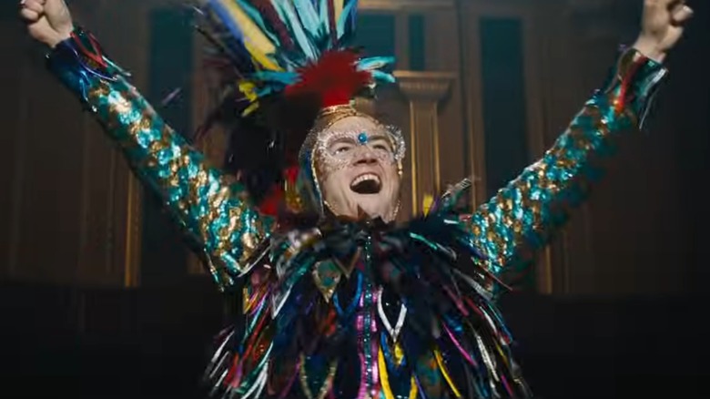 Elton John in colorful costume lifting arms yelling in still from Rocketman