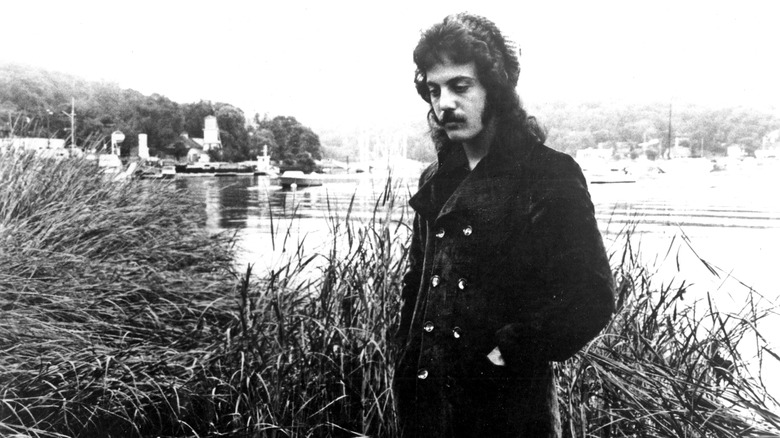 Billy Joel standing by river in long peacoat 1971