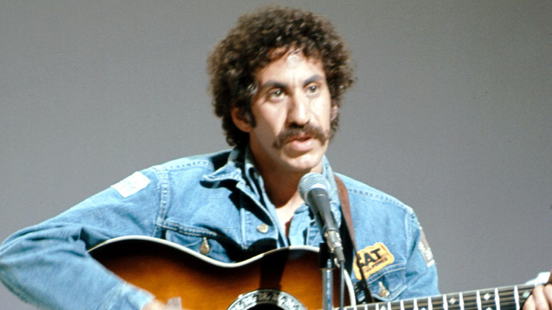 Jim Croce in denim jacket playing guitar and singing into mic on stage