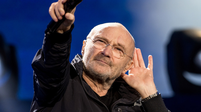 Phil Collins holding hand to ear and pointing mic at audience