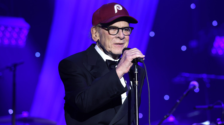 Art Garfunkel singing into the microphone wearing a burgundy baseball cap and tuxedo during a January 2026 performance