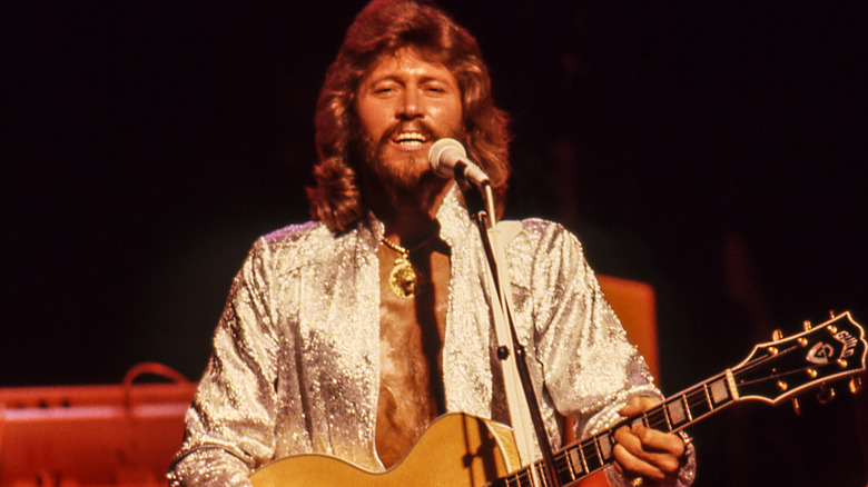 Barry Gibb performing with the Bee Gees on stage wearing a silver sparkly shirt and holding his guitar in 1979