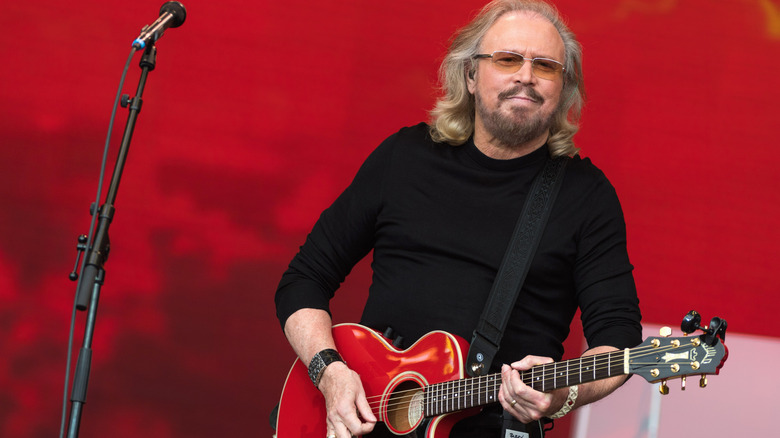 Barry Gibb playing a red guitar on stage in an all-black outfit and orange-tinted sunglasses