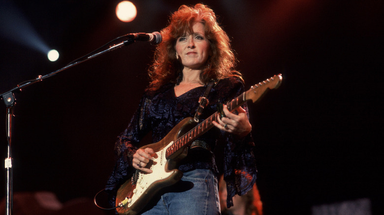 Bonnie Raitt playing the guitar onstage in a flowly navy top and blue jeans standing in front of a microphone