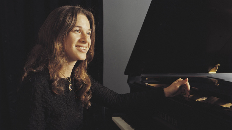 Carole King smiling and sitting at a piano