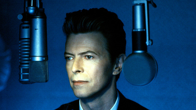 David Bowie in front of a blue background, with microphones hanging next to his head.
