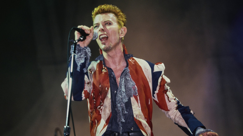 David Bowie on stage, singing into a microphone while gripping it with his right hand.