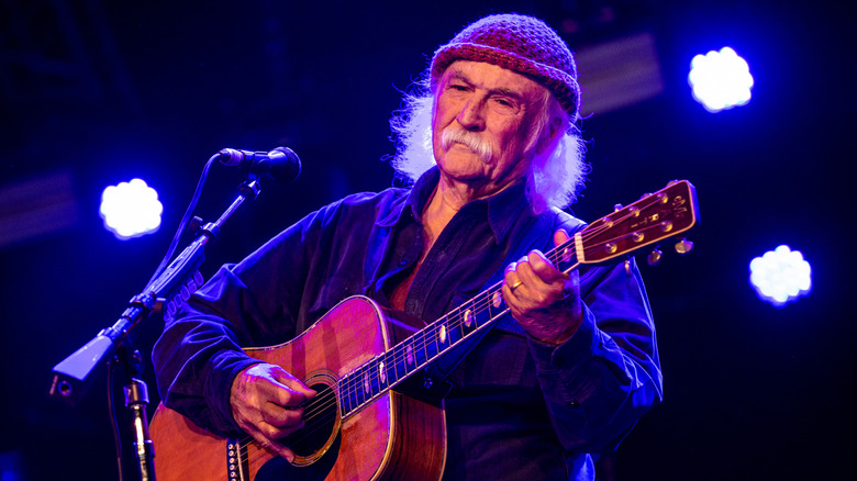 David Crosby performing on stage in Amsterdam in 2018 in a knit beanie with blue lights illuminating him from behind