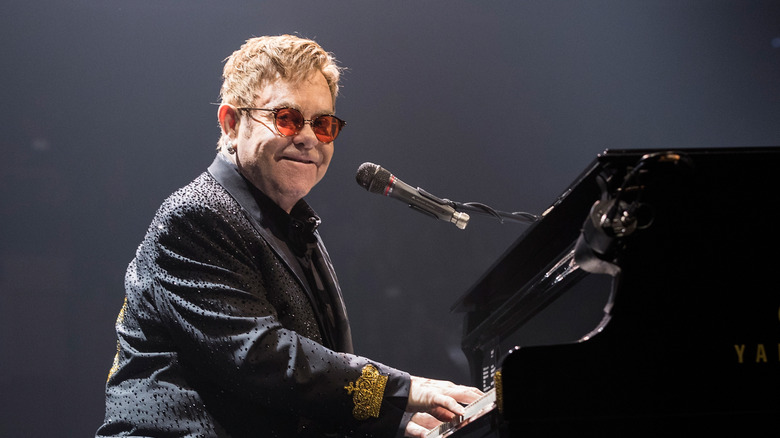 Elton John smiling and playing the piano on stage in red sunglasses