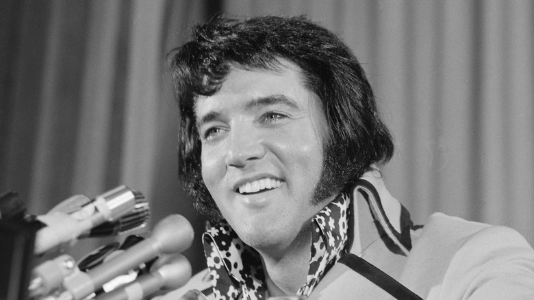 Elvis Presley smiling in front of multiple microphones at a press conference.
