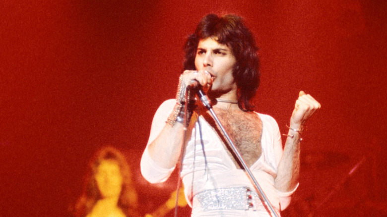 Freddie Mercury performing with Queen with long hair, wearing a white low cut bodysuit with a silver belt, silver arm cuff, and choker