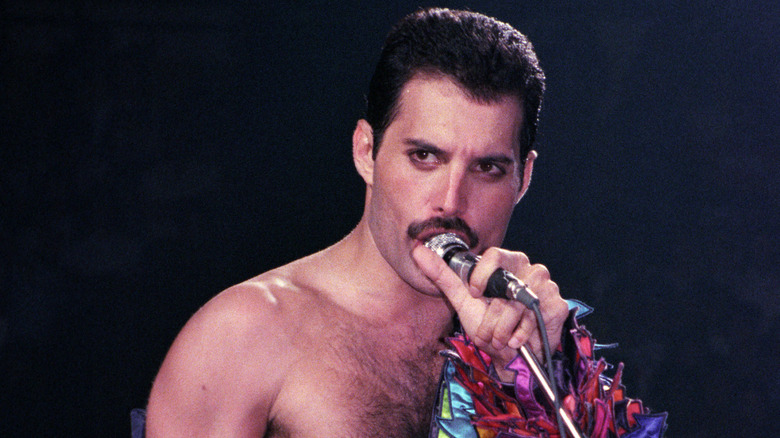 Freddie Mercury singing into a microphone with a bit of his colorful, mosaic visible on his left arm during a performance in 1983