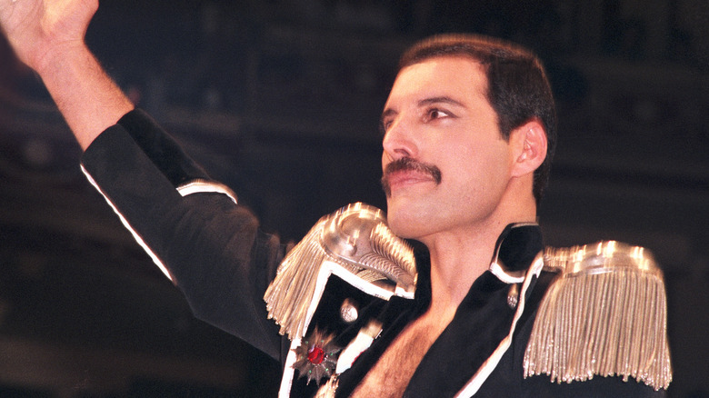 Freddie Mercury in a military style jacket with golden shoulder tassels raising his arm to a crowd during a 1985 concert