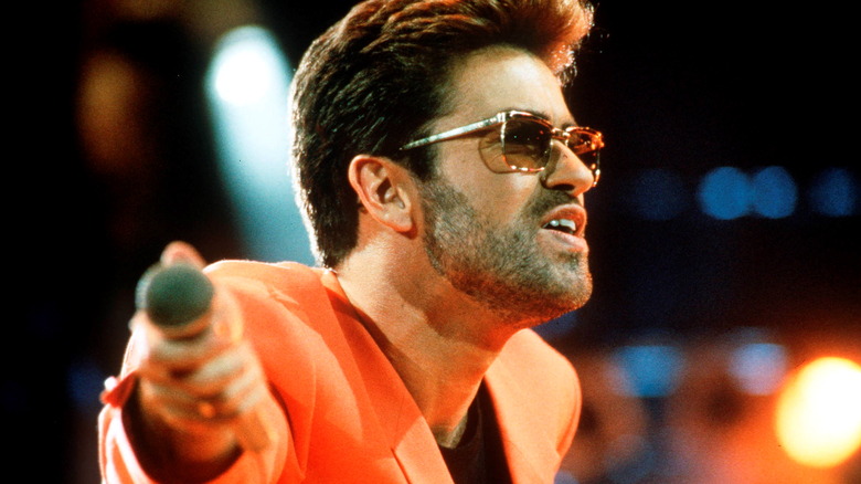 George Michael on stage holding out mic in orange jacket