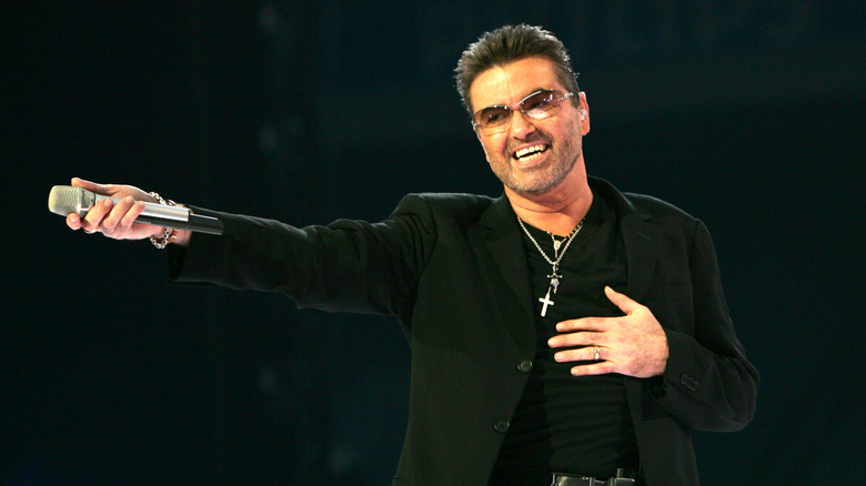 George Michael smiling in black suit on stage holding his microphone out