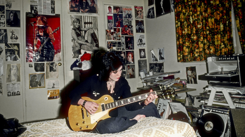 Joan Jett sat on a bed playing a guitar.