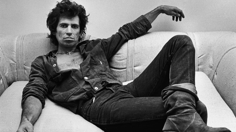 Black and white photo of Rolling Stones' guitarist Keith Richards lounging on a couch in a dark button up shirt, jeans, and boots in the 1980s