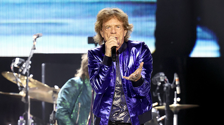 Mick Jagger performs on the final night of the Hackney Diamonds tour in 2024, extending a hand out to the audience in a purple metallic jacket and silver shining shirt
