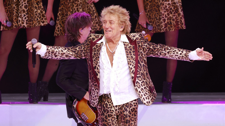 Rod Stewart holds a microphone on stage with his arms open while wearing a cheetah print suit